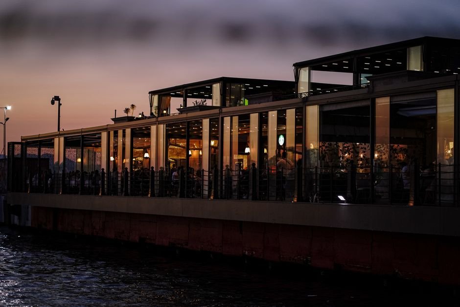 A modern restaurant by the water at twilight with warm interior lighting and sunset sky.